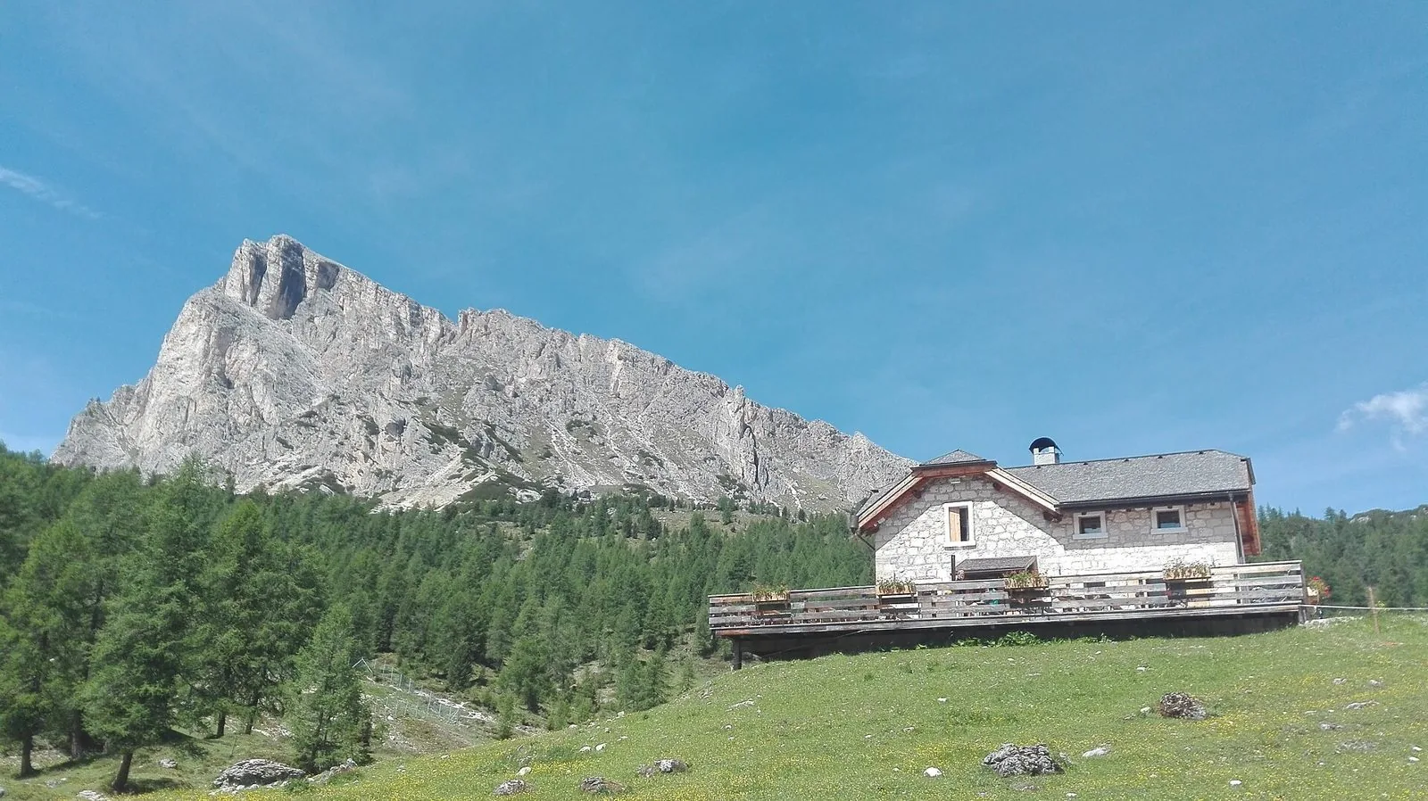 Malga Giau a San Vito di Cadore, edificio in pietra con terrazza in legno sui pascoli, bosco di larici e parete dolomitica sullo sfondo